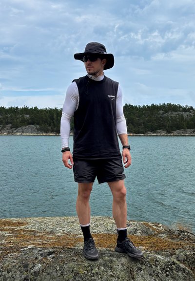 Camisa sin mangas negra sobre una camiseta de manga larga blanca, pantalones cortos negros, un sombrero de ala ancha, gafas de sol y calzado oscuro junto a un cuerpo de agua.