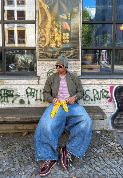 Joven con vaqueros de gran tamaño, chaqueta verde y gorro sentado en un banco de madera contra una pared cubierta de grafitis con un cartel de sushi detrás.