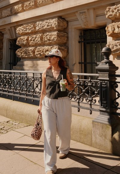 Top negro sin mangas, pantalones de pierna ancha a rayas y una gorra blanca. Sostiene una bebida y un bolso de leopardo, caminando junto a un edificio de piedra.