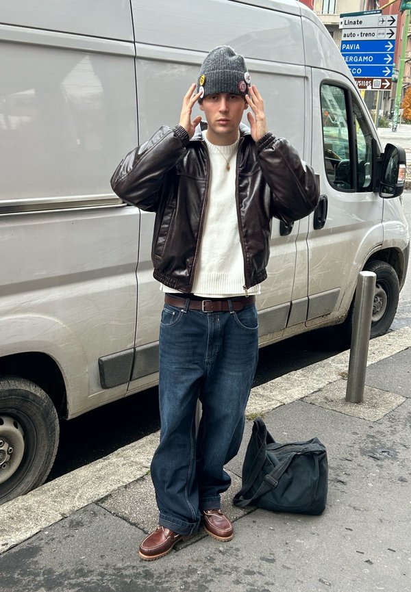 Ung mann iført grå strikkelue, brun skinnjakke, hvit genser, blå jeans og brune støvler, stående ved en hvit varebil på en bygate.