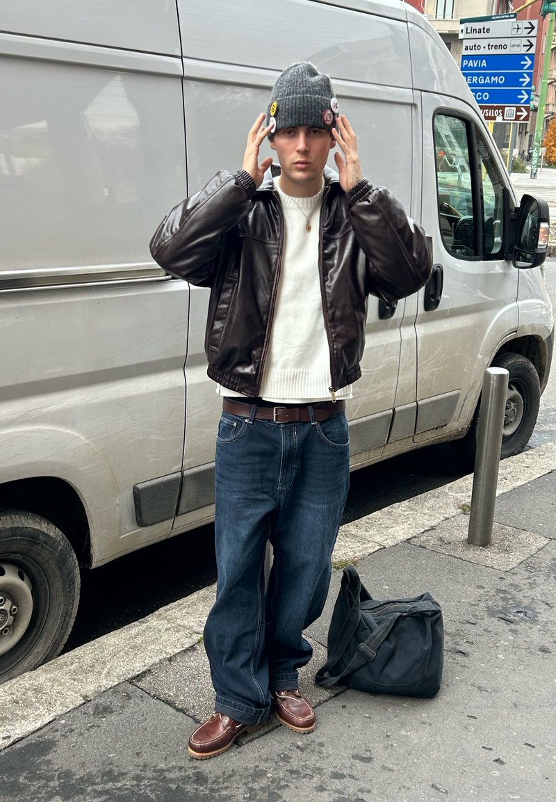 Ung mand iført grå strikhue, brun læderjakke, hvid sweater, blå jeans og brune støvler, stående ved siden af en hvid varevogn på en bygade.