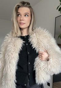 Étole en fausse fourrure de couleur crème, texture douce avec de longs poils ébouriffés, portée sur un haut noir boutonné, avec un col rond et des boutons argentés.