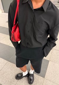 Pessoa vestindo camisa preta abotoada e calções, meias brancas, mocassins pretos, e carregando uma mala de couro vermelha ao ombro.