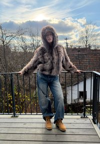 Brown faux fur cropped jacket with a hood, paired with loose blue denim jeans and tan boots, on a balcony with a cloudy sky background.