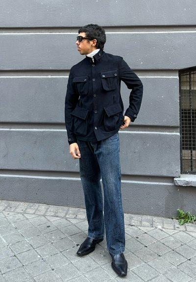 Hombre con chaqueta negra, cuello alto blanco, vaqueros azules y zapatos negros puntiagudos de pie en la acera contra una pared gris, con gafas de sol.