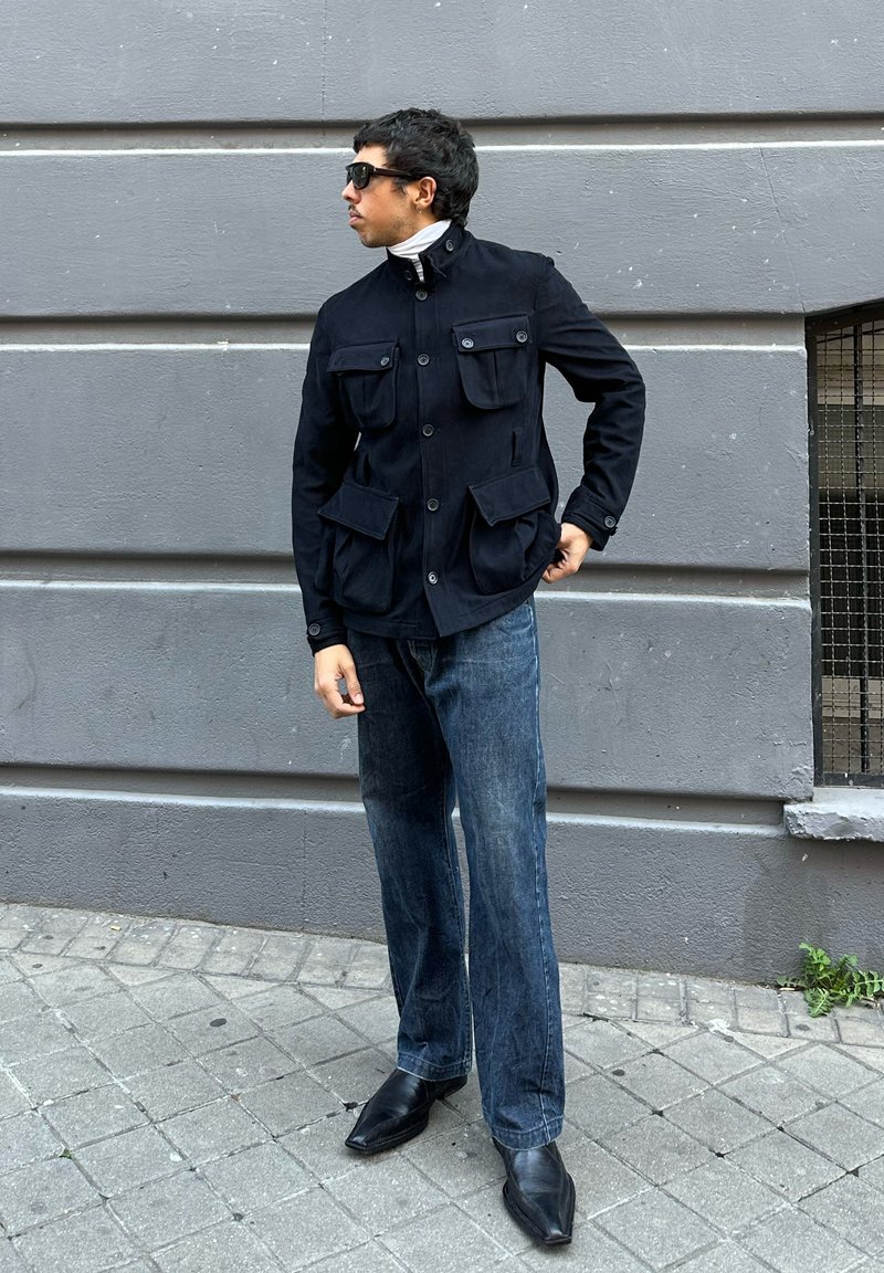 Άνδρας με μαύρο μπουφάν, λευκό ζιβάγκο, μπλε τζιν και μαύρα μυτερά παπούτσια που στέκεται στο πεζοδρόμιο κοντά σε γκρι τοίχο, φορώντας γυαλιά ηλίου.