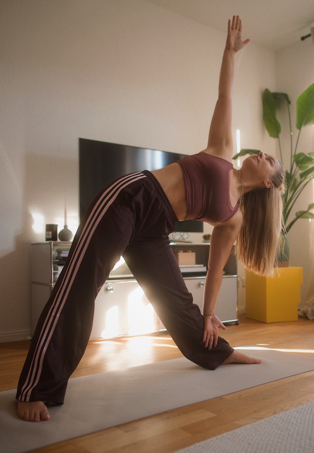 Uma pessoa com uma t-shirt de alças marrom-escura e calças de fato de treino escuras com riscas rosas executa uma pose de yoga num tapete cinza em uma sala bem iluminada.