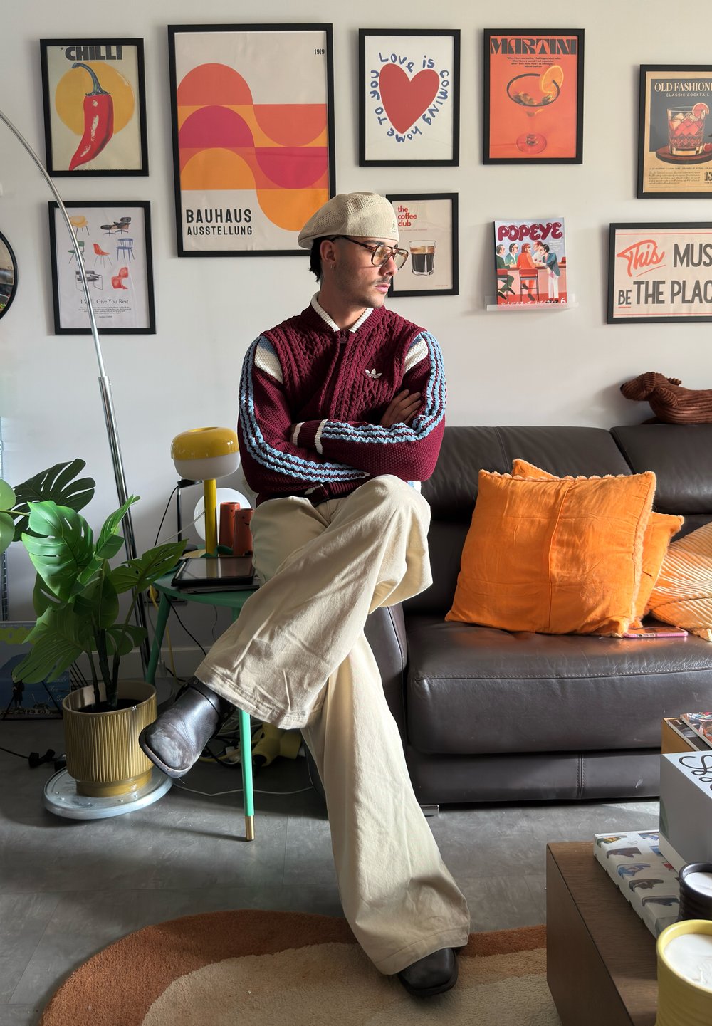 Vaatanpunane kootud džemper siniste ja valgete triipudega, beežid laiade säärtega püksid, mustad saapad ja hele müts. Moodne sisekujundus raamitud kunstiteostega.