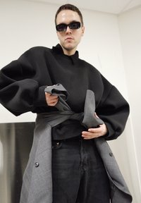 Sweat-shirt noir surdimensionné avec épaules tombantes, tissu gris enroulé autour de la taille, et pantalon en denim noir. Des lunettes de soleil complètent le look.