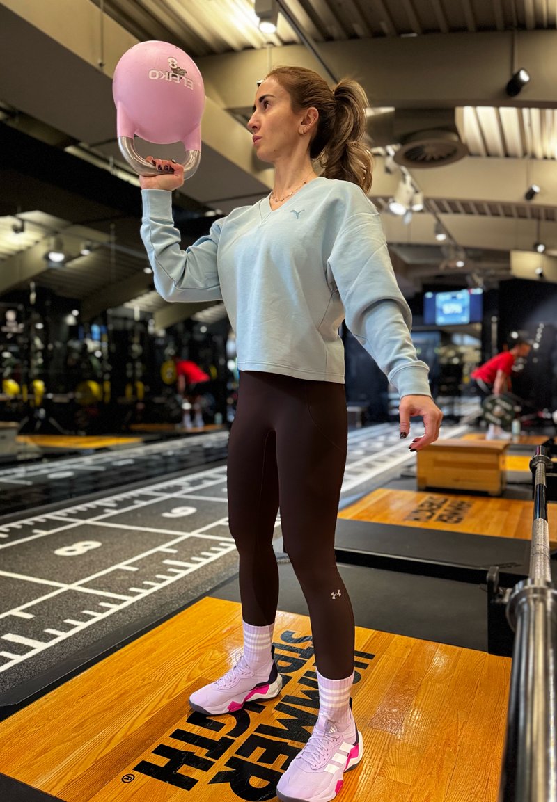 Ženska v telovadnici dviguje roza kettlebell z eno roko, stoji na lesenem odru, v ozadju vidna telovadna oprema in vadbeni prostor.