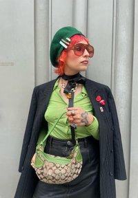 Boinas verdes, óculos de sol castanhos oversized, camisa verde de botão, blazer preto, saia de couro e uma bolsa estampada em castanho e verde.