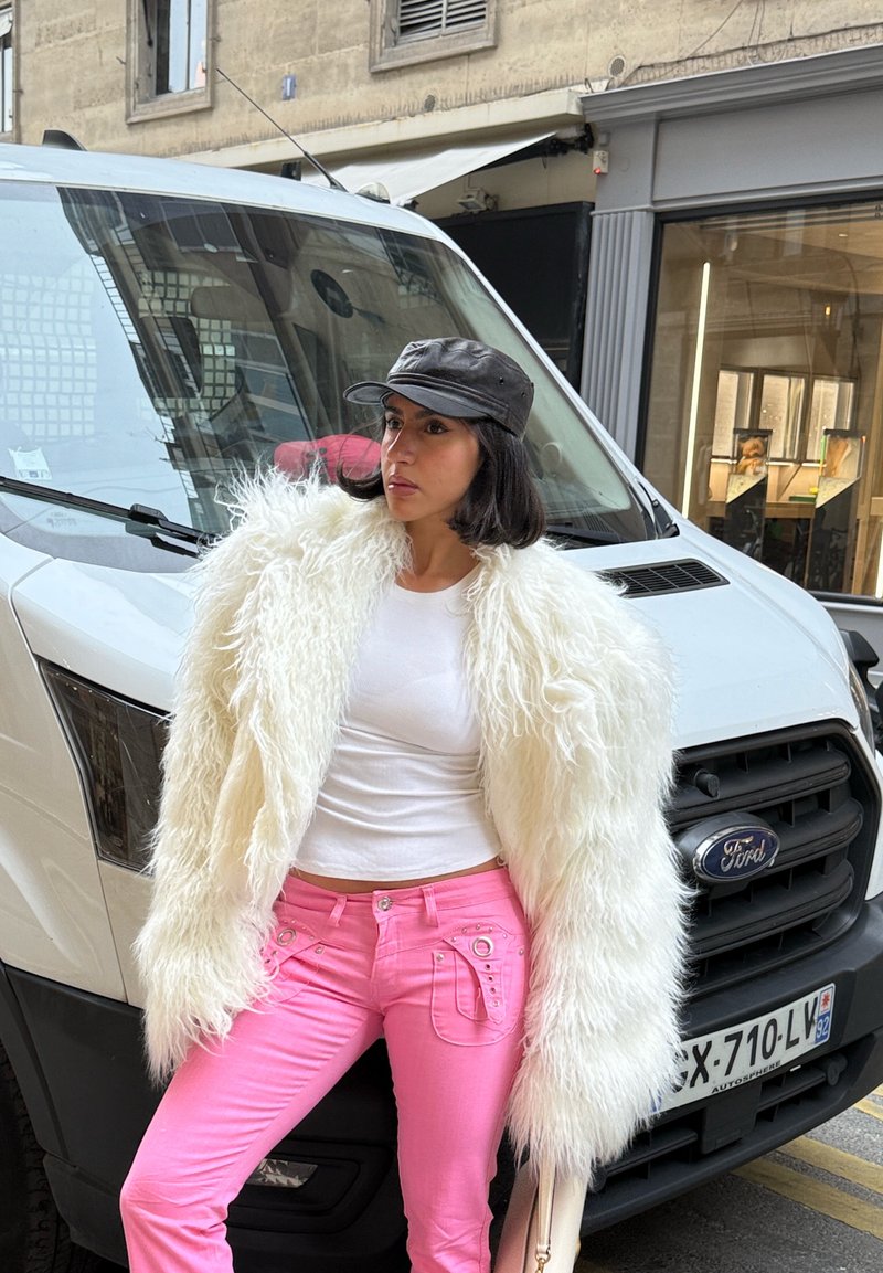 Frau in weißer Pelzjacke, weißem Oberteil, pinker Hose und schwarzer Kappe lehnt an einem weißen Ford-Van auf einer Stadtstraße.