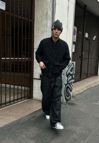 Jeune homme marchant sur un trottoir en ville, portant un bonnet gris avec des badges, une veste matelassée noire, un jean large foncé et des baskets blanches.