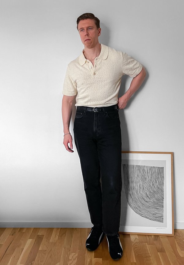 Mees valges lühikeste varrukatega polosärgis ja mustades teksades seisab valge seina ääres, kus puidust põranda peal on raamitud mustvalge abstraktne kunst.