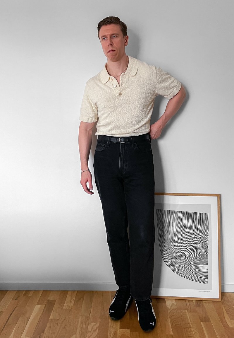 Homme en polo blanc à manches courtes et jean noir, debout contre un mur blanc avec une œuvre d'art abstraite en noir et blanc encadrée reposant sur un sol en bois.