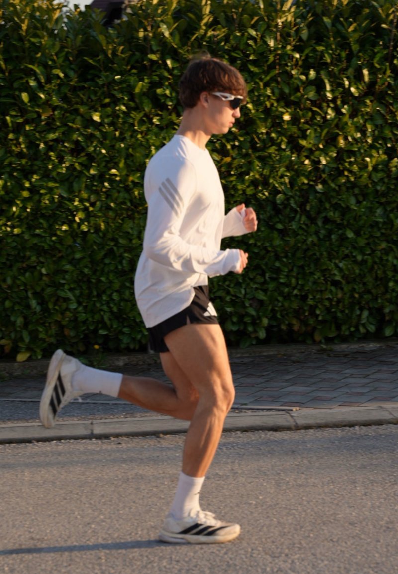 Jeune homme portant une chemise blanche à manches longues, un short noir et des lunettes de soleil, en train de jogger sur une route pavée avec une haie verte en arrière-plan.