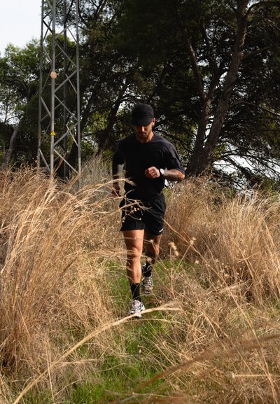 Hombre con ropa deportiva negra y gorra corriendo por un sendero estrecho entre hierba seca y alta, con árboles y una torre metálica al fondo.