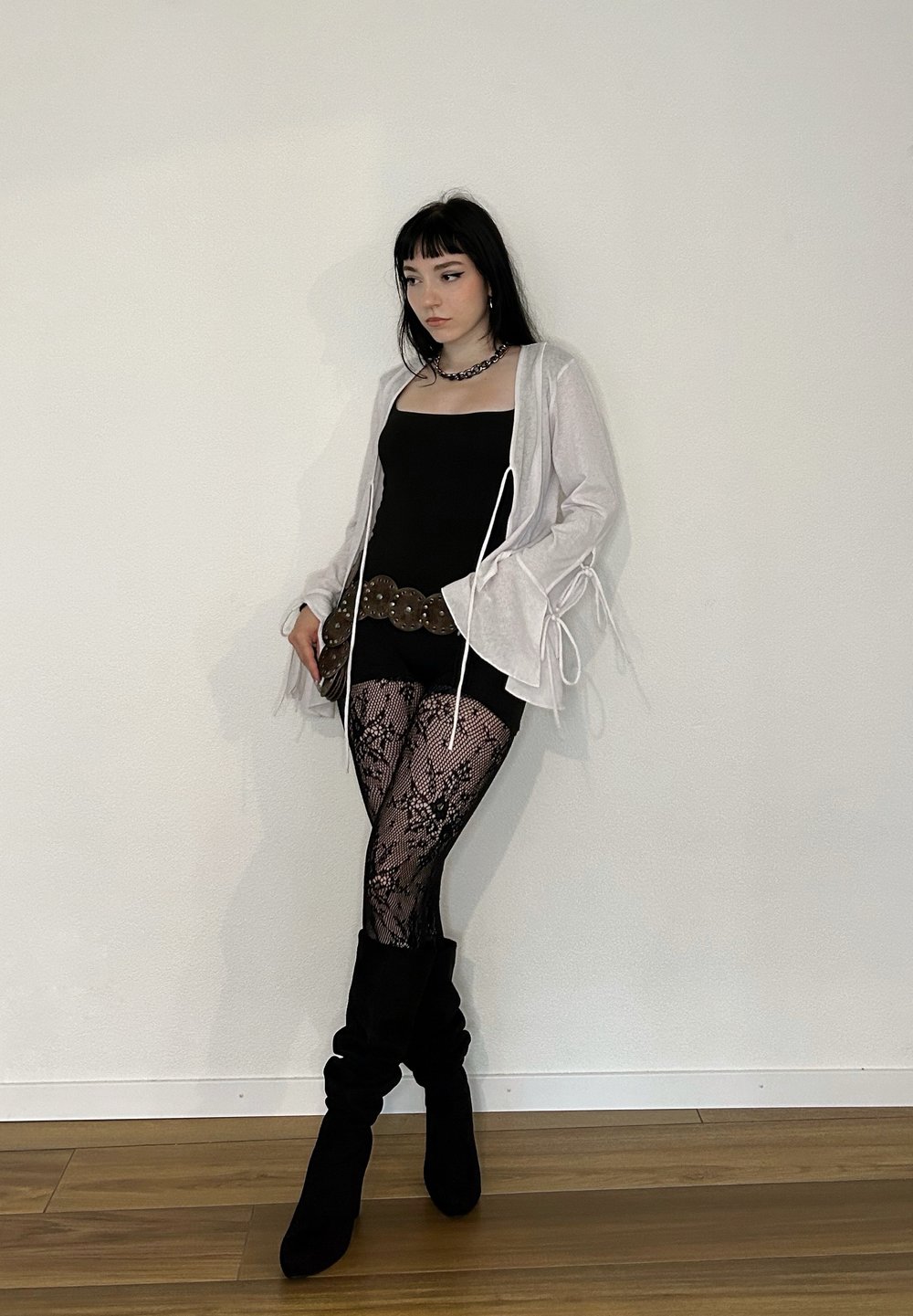 Fekete ujjatlan top, világosszürke kardigán megkötőkkel, fekete csipkés leggings, széles barna öv, és fekete térd fölé érő csizma fapadlón, fehér fal előtt.