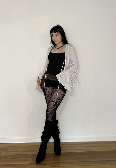Top negro sin mangas, cárdigan gris claro con lazos, leggings de encaje negro, cinturón ancho marrón y botas negras hasta la rodilla sobre un suelo de madera frente a una pared blanca.