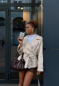 Kvindelig med krøllet hår i en beige trench coat, briller og guldøreringe, der sipper en drik gennem et sugerør, mens hun holder en mørkebrun håndtaske ved en glasdør.