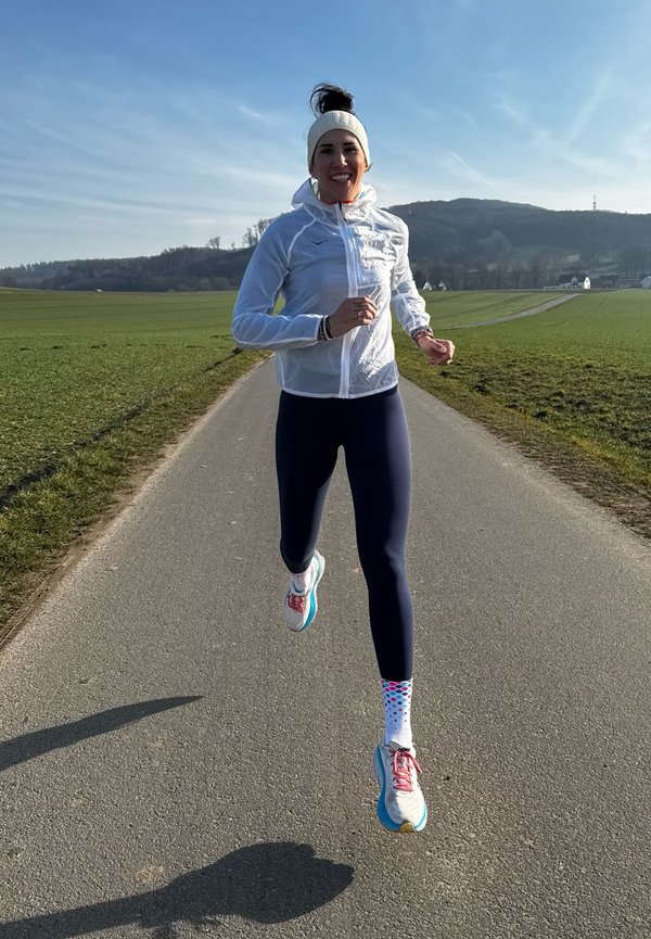 Runner in a light, transparent white jacket, navy leggings, multicolored polka dot socks, and blue running shoes on a pathway through grass.