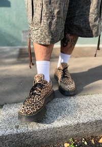 Buty w imitację skóry lamparta z teksturowaną powierzchnią, czarnymi podeszwami i czarnymi sznurówkami, noszone z białymi skarpetkami i szortami w wzory.