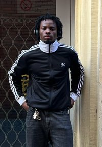 Casaco preto com fecho éclair, riscas brancas e logo da Adidas, combinado com jeans escuros. A usar auriculares pretos. Em pé contra uma parede.