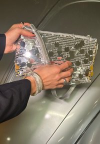 Srebrna torbica s sequinsom in prozornim materialom, z zadrgo in odstranljivo naramnico. Ročna torbica z vidnimi srebrnimi zapestnicami.