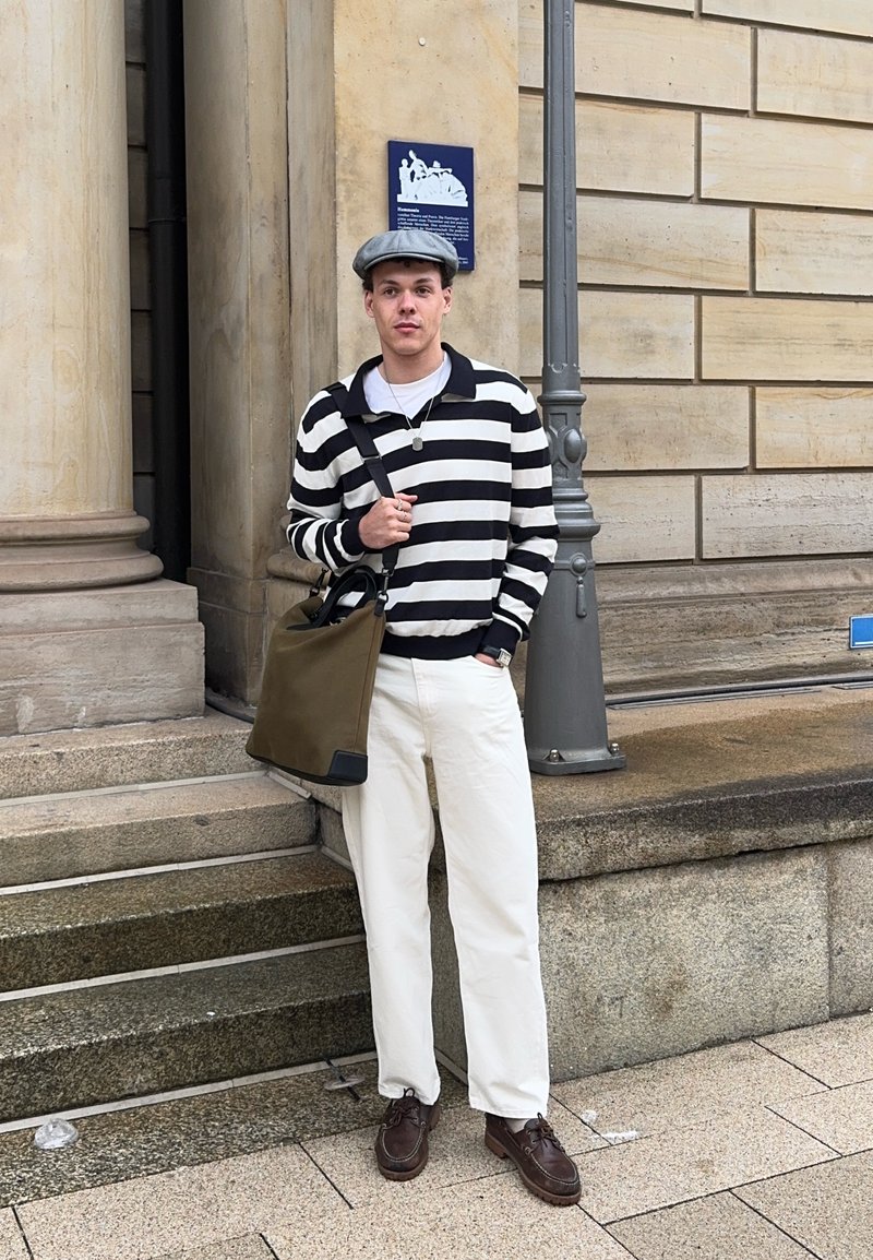 Young man wearing a striped jumper, white trousers, flat cap, and brown shoes, standing beside stone steps and a lamp post outdoors.