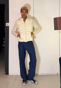 Νεαρό άτομο με ξανθά σγουρά μαλλιά, φορώντας κρεμ μπουφάν με ραφές, μπλε παντελόνι με σχέδια και ασπρόμαυρα αθλητικά παπούτσια, που στέκεται σε εσωτερικό χώρο.