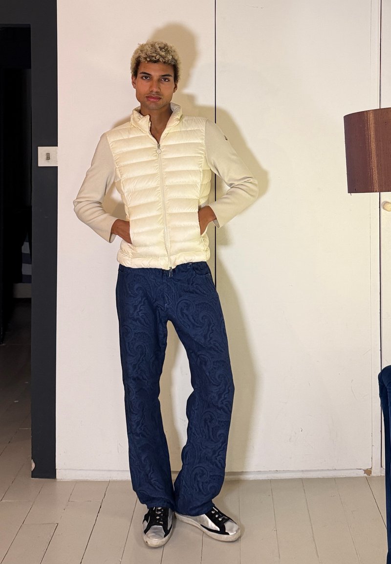 Jeune personne aux cheveux blonds bouclés portant une veste matelassée crème, un pantalon bleu à motifs et des baskets noires et blanches, debout à l'intérieur.