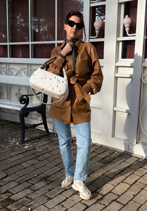 Una mujer con gafas de sol, una chaqueta de cuero marrón, vaqueros y zapatillas posa con un bolso blanco delante de un edificio.