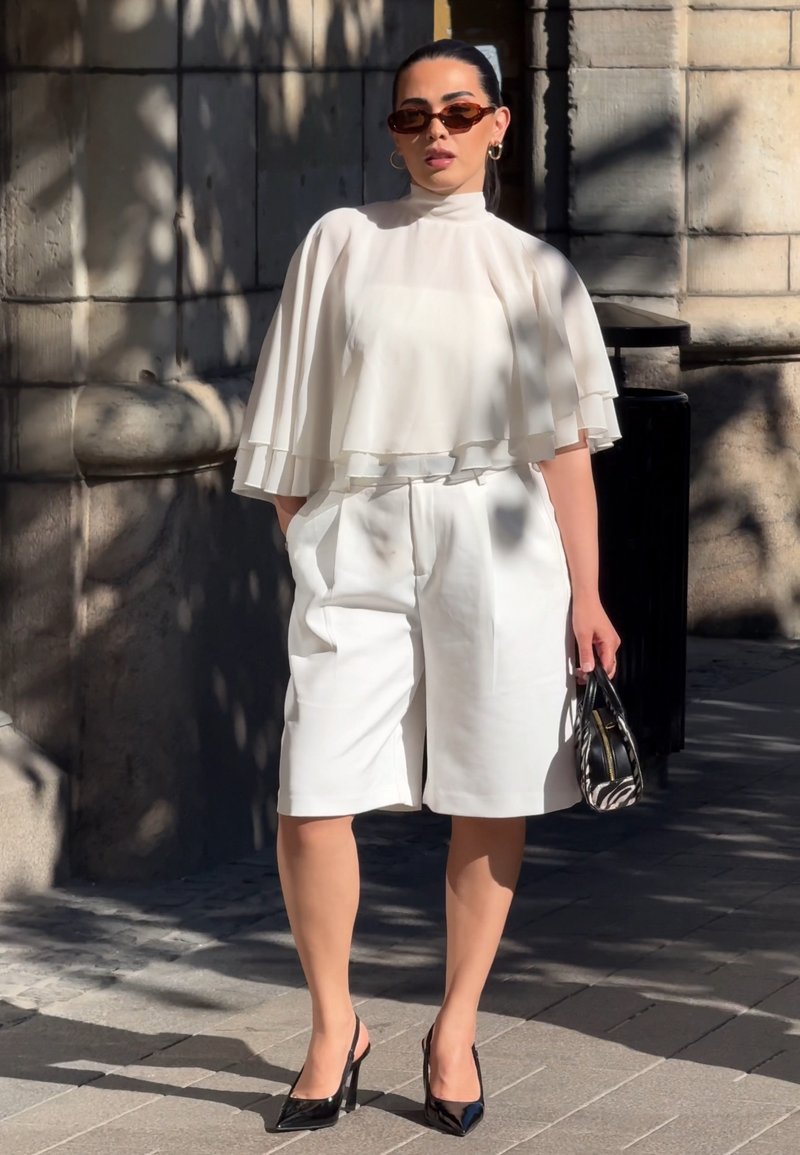 Femme debout à l'extérieur portant un haut cape blanc à plusieurs couches, un short blanc jusqu'aux genoux, des talons noirs, des lunettes de soleil et tenant un petit sac à main noir.