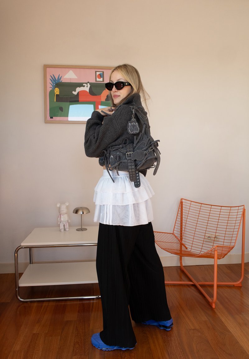 Femme portant des lunettes de soleil noires, une jupe blanche superposée et un pantalon noir, tenant un sac noir détaillé, debout dans une pièce avec une chaise en fil orange et une œuvre d'art.