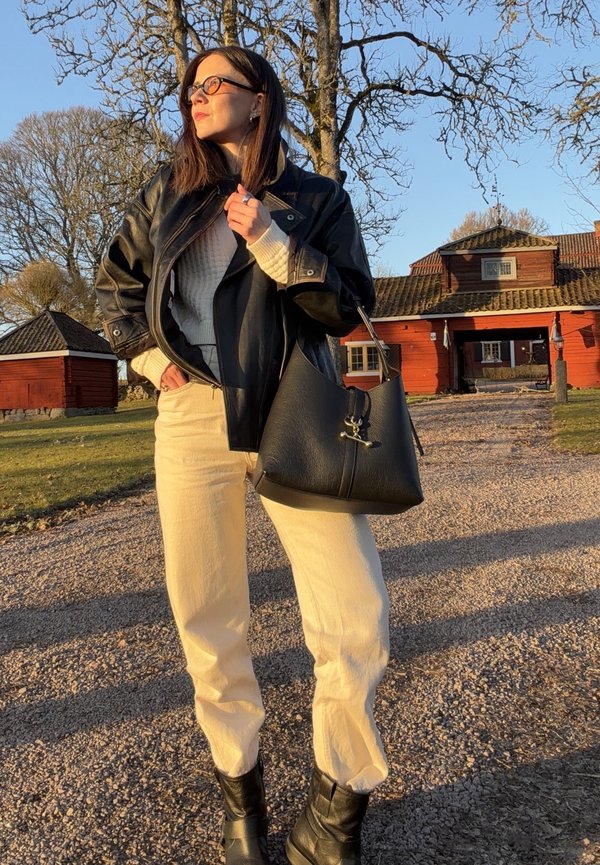 Femme portant une veste en cuir noire, un pull crème, un pantalon clair et des bottes noires, tenant un sac à main noir à l'extérieur près de bâtiments en bois rouges.