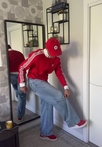 Chaqueta roja de Adidas con cremallera y rayas blancas, jeans de pierna ancha en azul claro y zapatillas rojas. Un espejo al fondo refleja el conjunto.