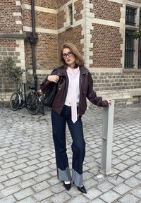 Brun skinnjacka, vit blus med rosa slips, mörka jeans med kontrasterande fåll, svarta högklackade skor och svart flätad väska. Bakgrund med kullersten.