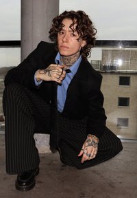 Pantaloni negri cu dungi subțiri, cămașă albastră și sacou negru. Persoană cu păr creț și tatuaje vizibile, îngenuncheată lângă o fereastră mare.