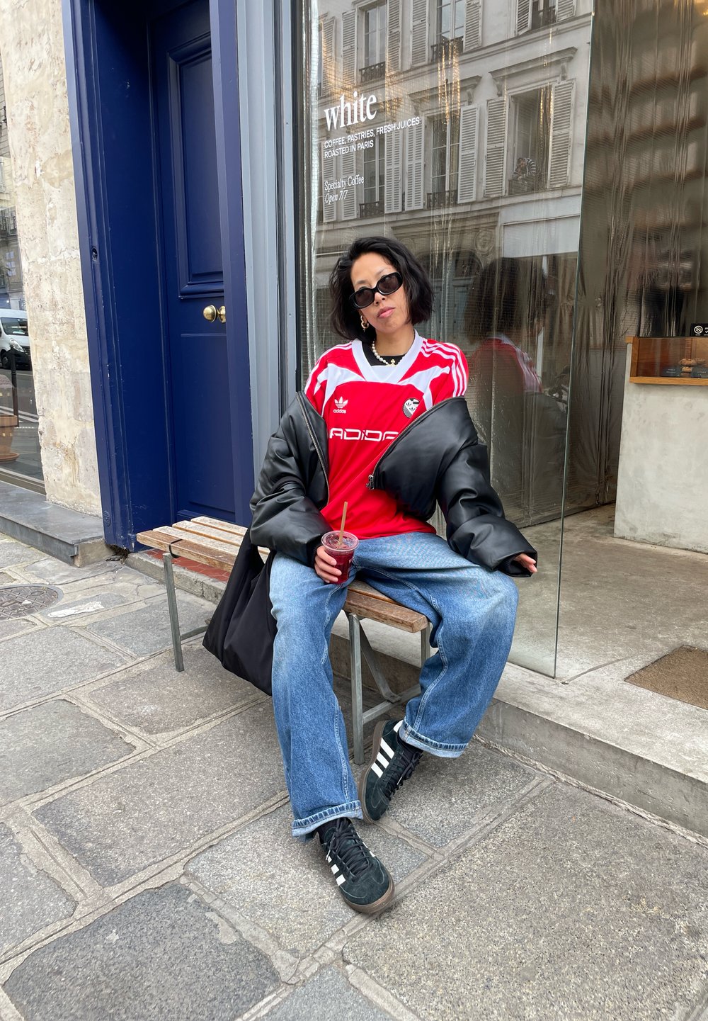 Rood Adidas-shirt met witte strepen, zwarte oversized jas, blauwe jeans en zwarte sneakers met witte strepen. Houdt een rode drank vast.