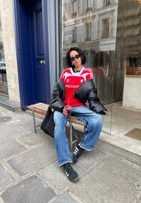 Rood Adidas-shirt met witte strepen, zwarte oversized jas, blauwe jeans en zwarte sneakers met witte strepen. Houdt een rode drank vast.