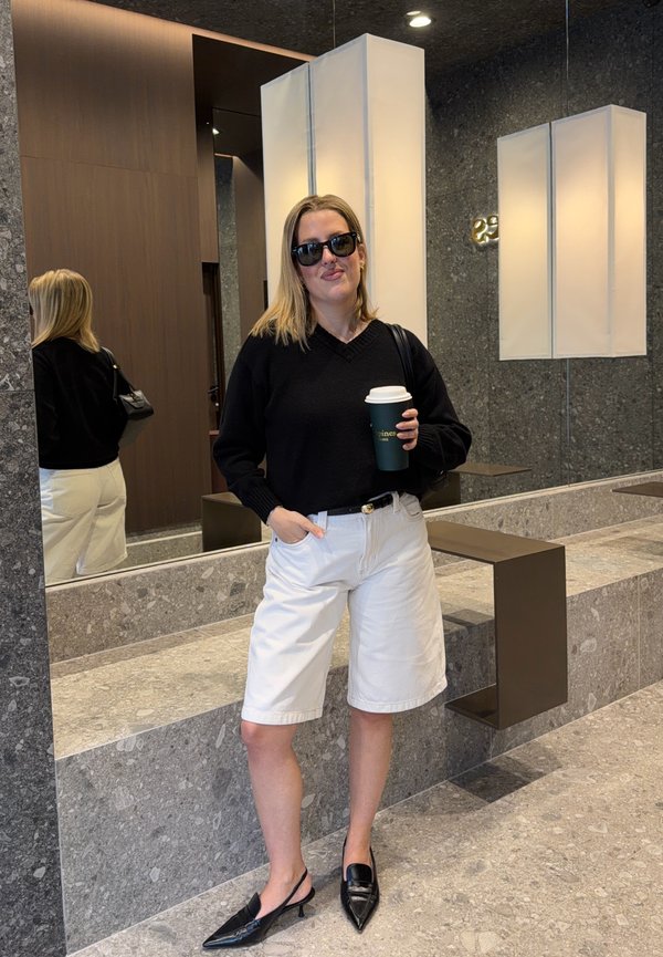 Kvinde i sort sweater, hvide knælange shorts, sorte spidse sko og solbriller, der holder en kaffekop og står i et moderne sten- og træinteriør.