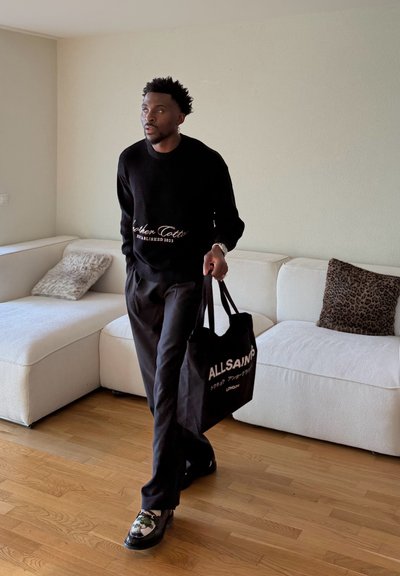 Hombre con suéter y pantalones negros camina dentro de una habitación sosteniendo una bolsa tote negra de AllSaints, con un sofá blanco y cojines con estampado animal al fondo.