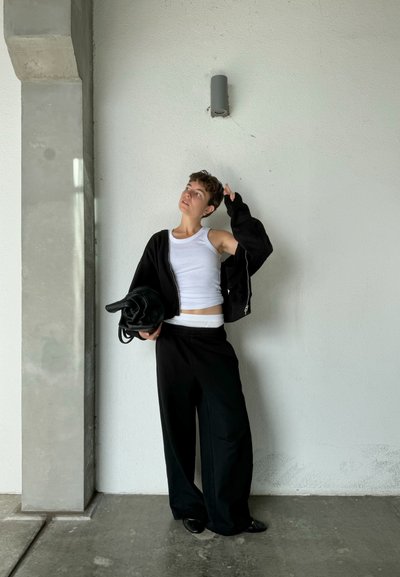 Sudadera negra con cremallera, camiseta sin mangas blanca y pantalones negros holgados. La modelo, que sostiene un bolso negro, se encuentra de pie junto a una pared blanca y un pilar de concreto.