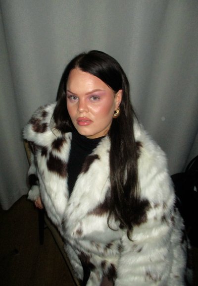 Abrigo de piel sintética blanca y marrón con una textura suave y un patrón de manchas, llevado sobre una blusa negra, complementado con pendientes de aro dorados.