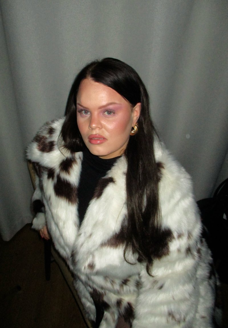 White and brown faux fur coat with a soft texture and spotted pattern, worn over a black top, complemented by gold hoop earrings.