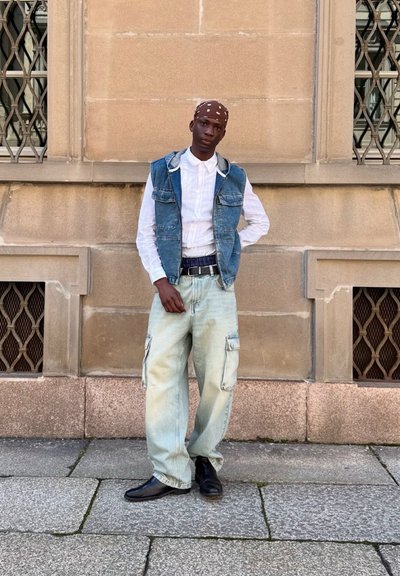 Chaleco de mezclilla sobre una camisa blanca, pantalones cargo holgados de color azul claro y zapatos negros, frente a una pared de piedra y una reja en la ventana.