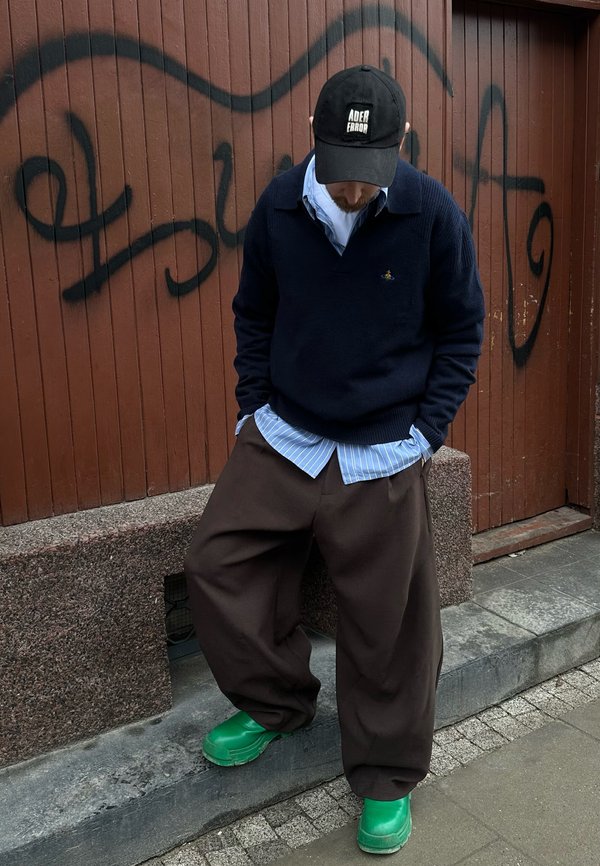 Man wearing a black cap, navy jumper over a blue striped shirt, wide brown trousers, and bright green shoes, standing on a city pavement next to graffiti.