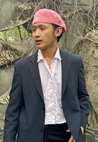 Black blazer over a light pink shirt, wearing a pink beret. Natural background with stone texture and vines. Neutral lighting.