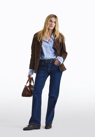 Mujer con chaqueta de ante marrón, camisa azul con botones, vaqueros azul oscuro, zapatos negros, sosteniendo un bolso de cuero marrón, de pie contra un fondo blanco.