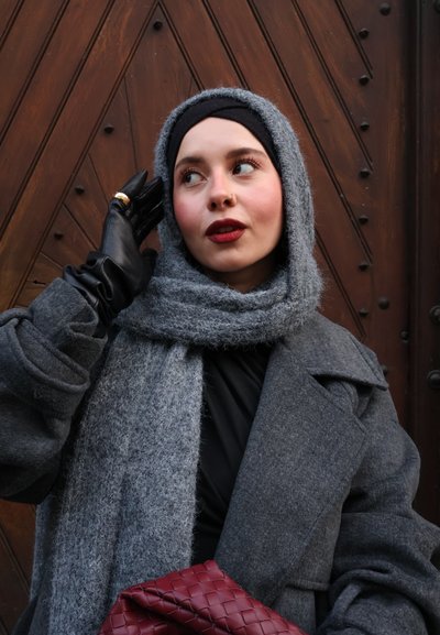 Abrigo gris con solapas grandes, combinado con una bufanda gris suave. Guantes negros, parte superior oscura y un bolso burdeos texturizado. Fondo con patrón de madera.